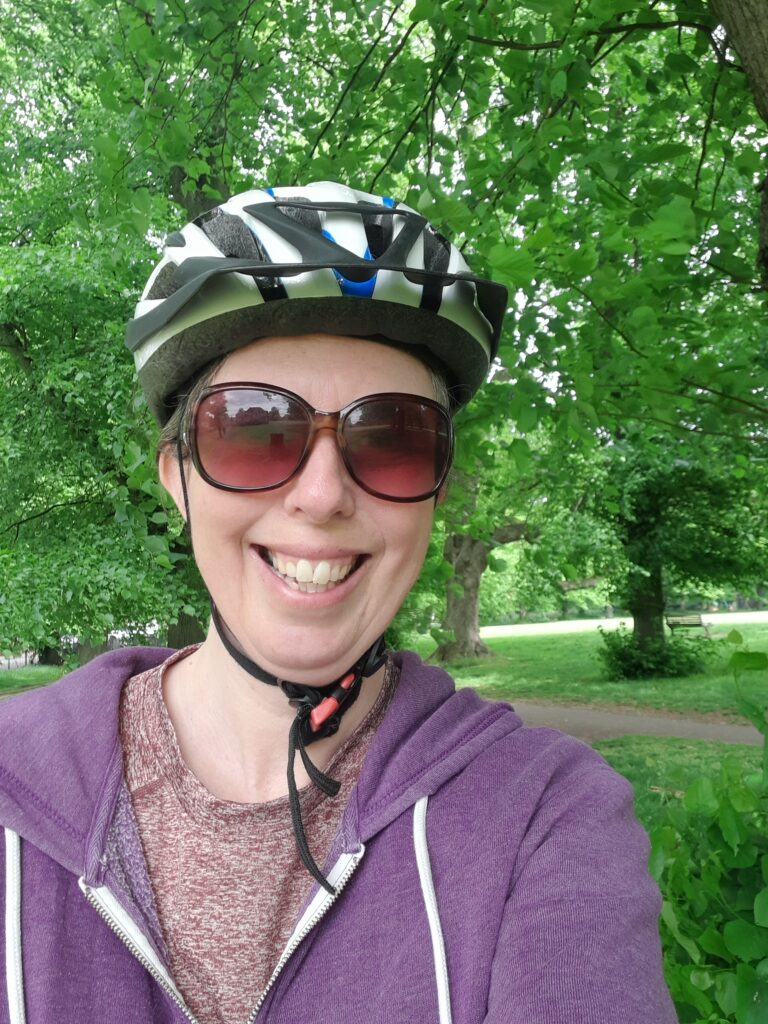 Sarah, a woman with light skin tone, wearing a a bicycle helmet and large sunglasses. Behind her are lots of trees and a path through them. She is smiling broadly.