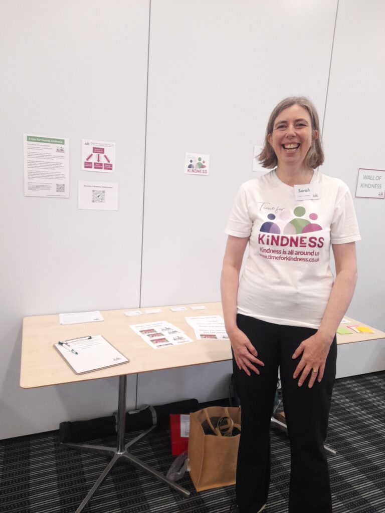 Sarah, a woman with light skin tone and shoulder length light brown/dark blonde hair wearing a white t-shirt with the Time for Kindness logo on it. She is smiling broadly and standing in front of a table on which are some papers and a clipboard. On the wall behind her are some A4 posters and the Time for Kindness logo.