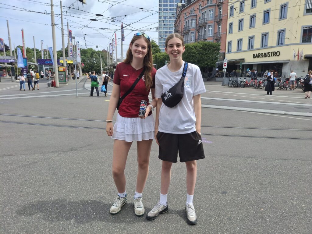 Two young women standing together on a plaza. Both have light skin tone and long brown hair, one wearing it loose, the other tied back. One is holding a can of Pepsi, the other is wearing a black cross body bag.