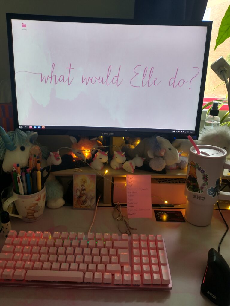a desk with a pink mat on it, pink keyboard, black ergonomic mouse, a monitor on a raised platform with pink desktop wallpaper reading “what would Elle do?” and various unicorns around the raised platform. Also on the desk are glasses, a tarot card, a large pink reusable bottle and some notes.