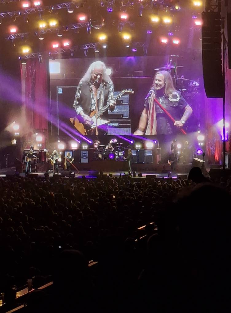 Wembley Arena filled with people standing in front of a stage on which Lynyrd Skynyrd are performing, a huge screen behind the stage showing them close up.