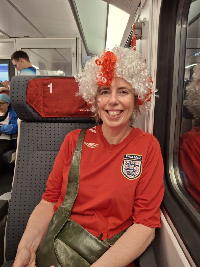 Sarah, a woman with light skin tone wearing a wig of white hair with a red cross on it and a red England Women’s football shirt, sat on a train. She is smiling broadly with an England flag painted on her cheek.