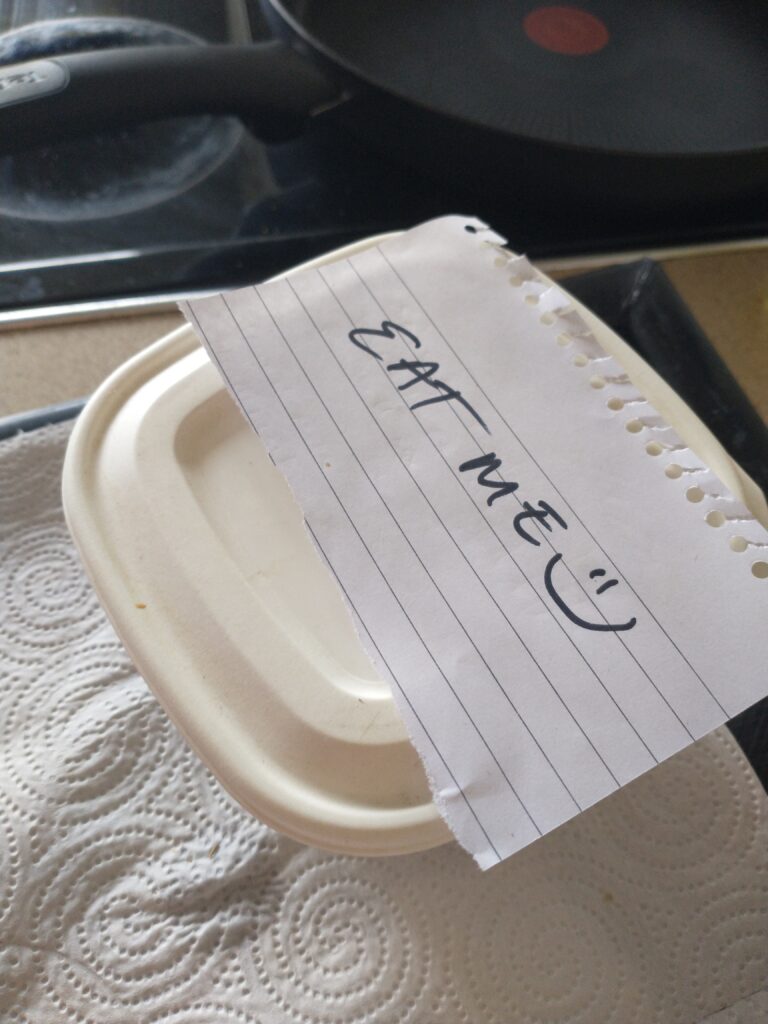 A plastic tub with a piece of lined paper resting on top which reads “eat me” with a smiley face.