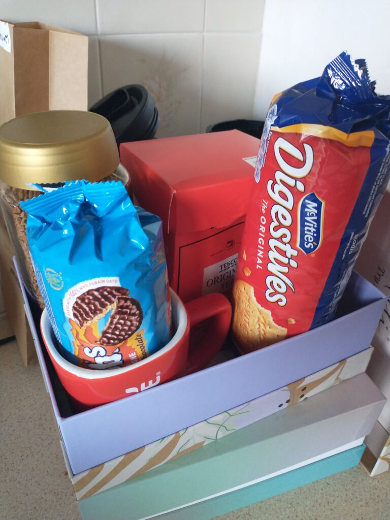 A pastel green box with another decorative box on top which is open. In the top box is a pack of Digestive biscuits, some Hobnobs, a box of tea bags, a jar of instant coffee and a red mug.