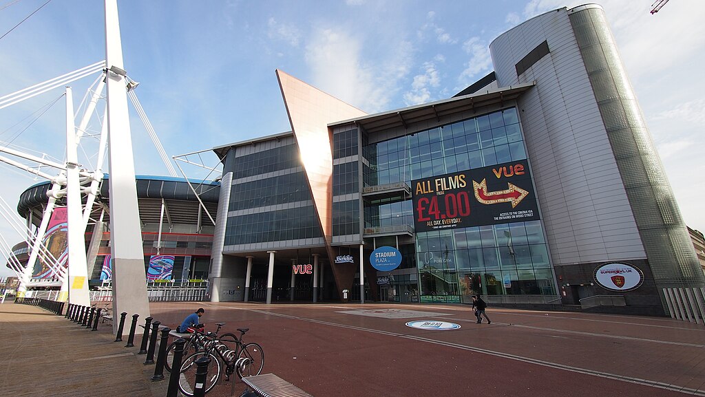 A large glass building with the Vue sign above one door and a large black sign reading “all films £4, Vue” with an arrow around the side of the building. In front of the building is a large pedestrian area, behind it is a stadium.
