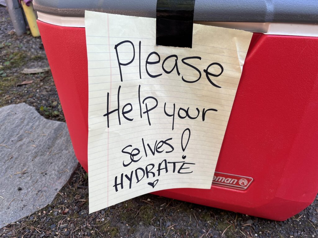 Bright red water cooler on some gravel with a note attached to it that reads “please help yourselves. Hydrate. ❤️”