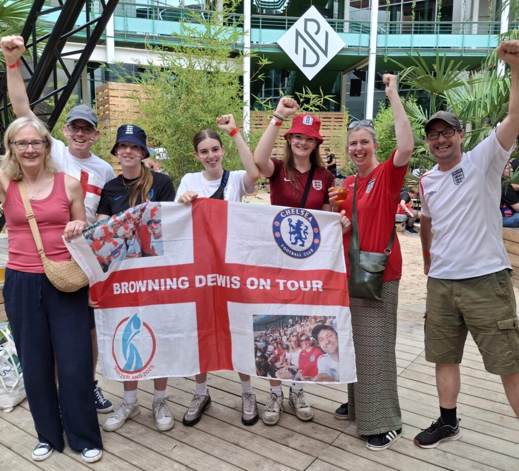A group of seven people stood in a line all holding a large England flag with “Browning Dewis on tour” written across the red cross, sports logos in two corners and photos of the group at other football events in the other corners. All of the members of the group are raising one hand in a fist and looking very excited.