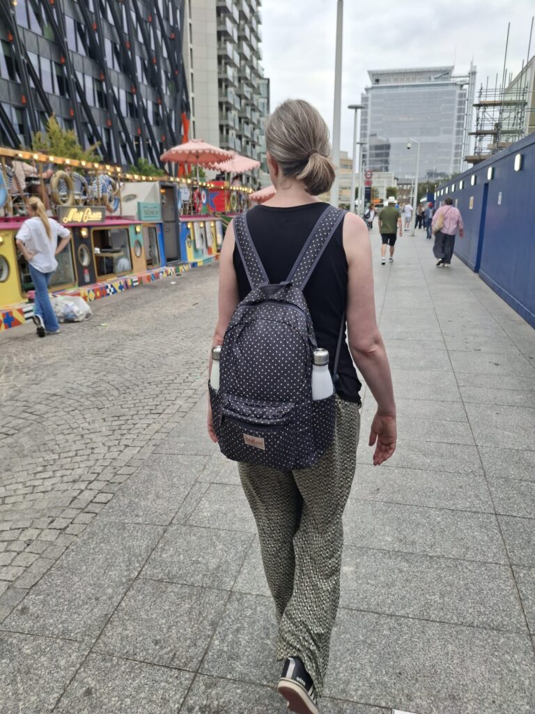 Sarah, a woman with light skin tone and long brown hair tied back, walking away from the camera down a London street. On her back is a dark blue Cat Kidston bag with white spots on it, there are reusable water bottles in both the pockets each side of the bag.