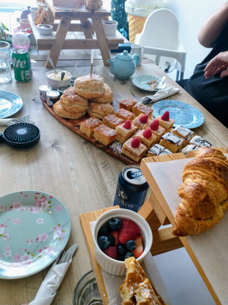 a table filled with all kinds of pastries. Many are on a large wooden board with scones and mini cakes, some on small wooden picnic tables which are sitting on the table alongside some containers of fruit. It all looks delicious.