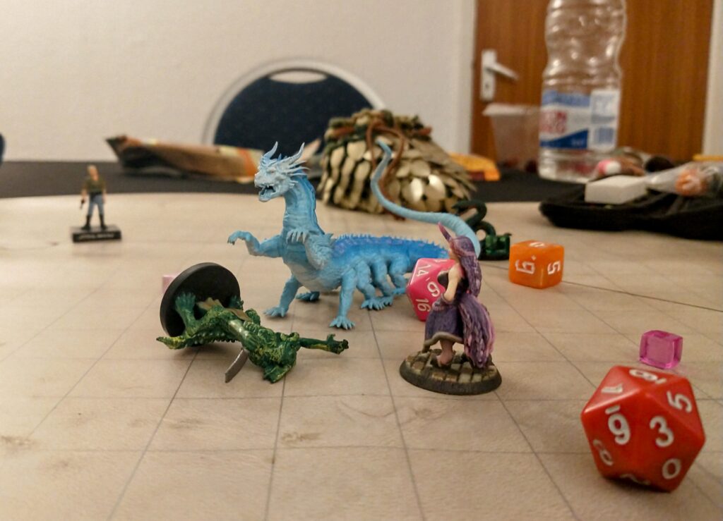 a beige dry erase mat with squares on it on a table. In the foreground is a red d20. In the centre a horse legged, human torsoed being with a unicorn horn on its head and pink and purple hair and tail, standing beside a green lizard creature which is laying on the floor. Next to them both is a large blue wingless dragon looking monster with 10 limbs and a long tail. 8 limbs act as legs and 2 act as short arms. It is roaring and looks vicious. Next to it is a pink d20. In the background out of focus is an elf shaped mini, an orange d6, and a green snake.