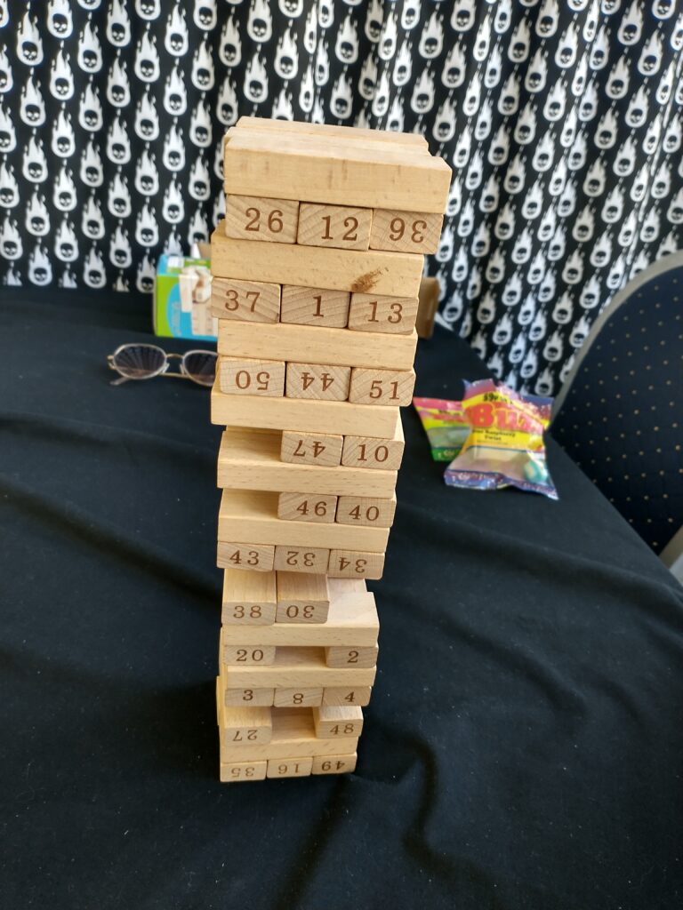 A Jenga tower of light brown wooden blocks with numbers on. The tower has a solid base but many pieces missing as it goes further up with a point about a third of the way up seemingly precariously balanced on just one centre block. The tower is on a table with a black cloth on it. There are snacks and a pair of glasses on the table behind it.