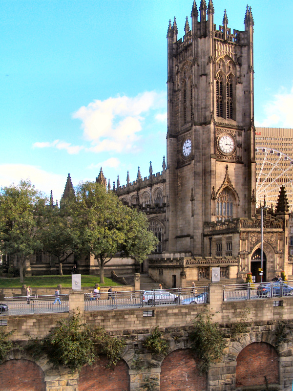 View of Manchester Cathedral and its grounds from a further away road. A blue sky with fluffy white clouds behind it, a road in front which people are driving and walking down.