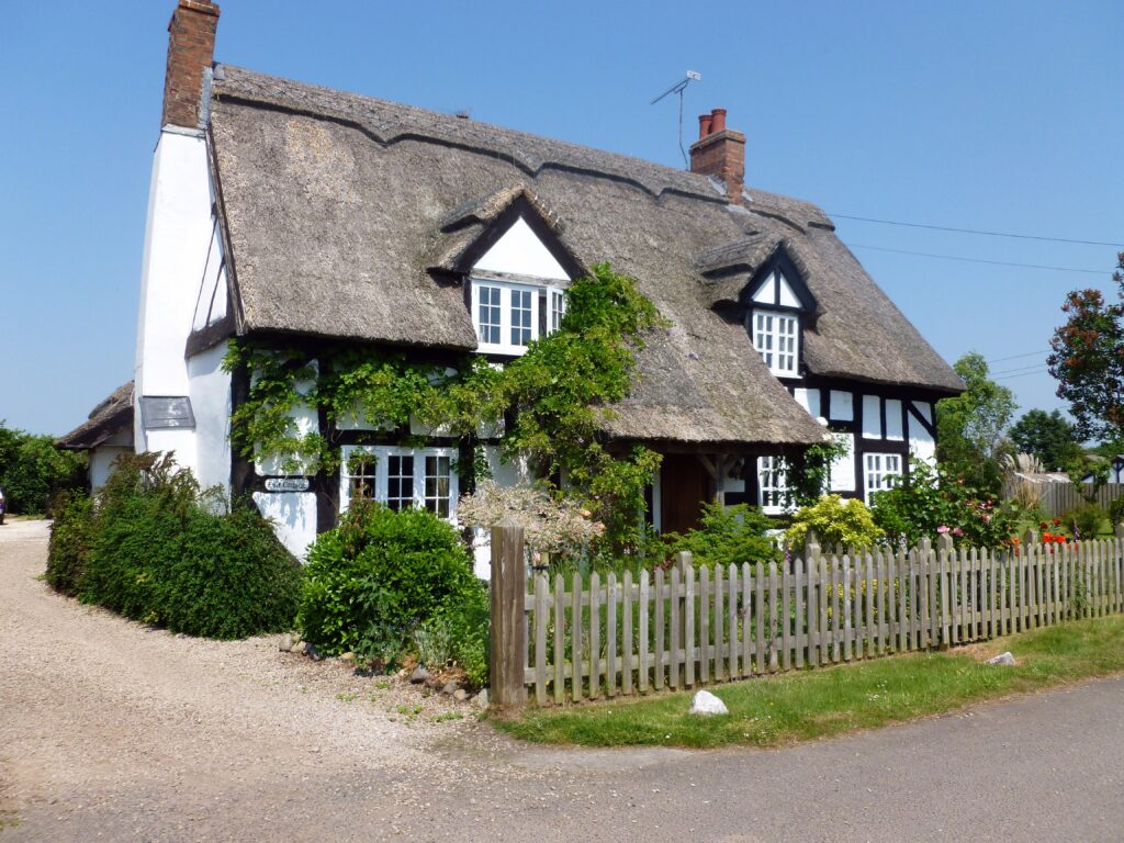 a village cottage in traditional English style with white walls, black beams and thatched roof. The sky above is pure blue and the cottage is surrounded by lush bushes and grass.