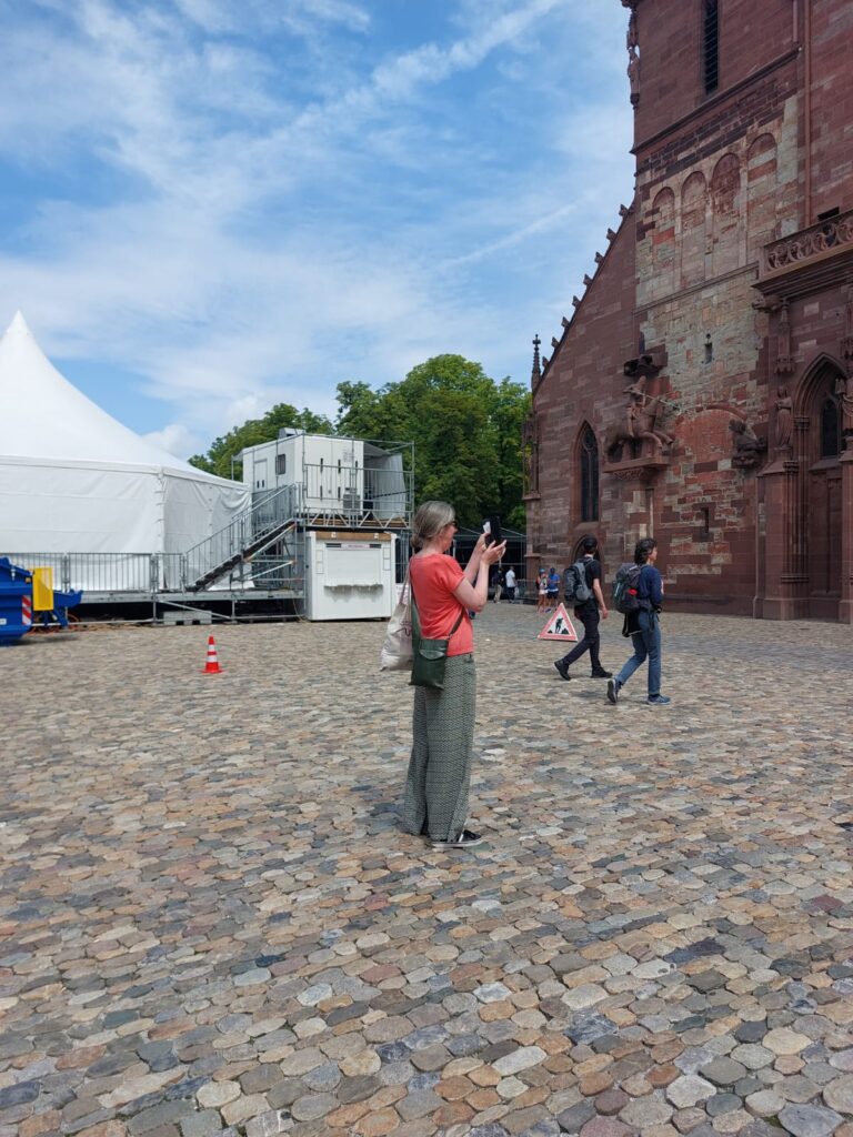 Sarah, a woman with light skin tone and long grey hair tied back, standing taking a photo in the plaza outside Basel Cathedral. Some other people are walking around in the area.