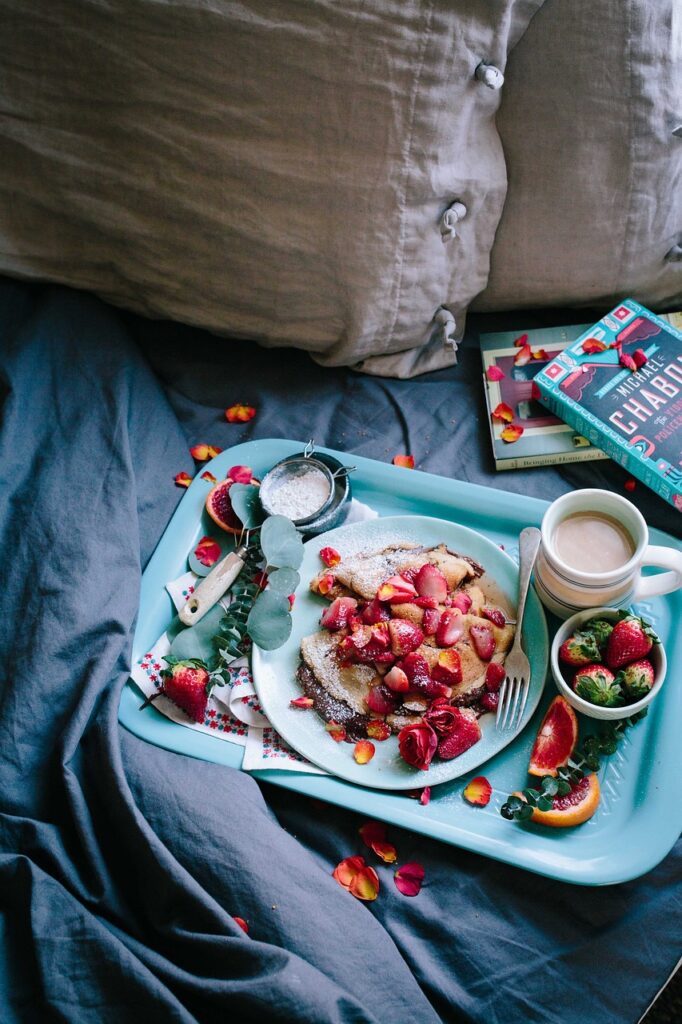 a blue tray with a plate of toast and strawberries, a coffee, some slices of orange, extra strawberries and cutlery on it. The tray sits on a roughly made bed with some books beside it.