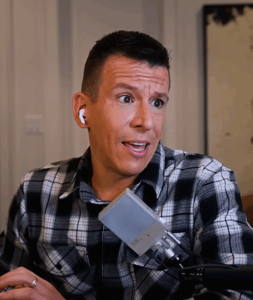Philip DeFranco, a man with light skin tone and short black hair with an animated face. He appears to in the process of saying something, a mic in front of him. He is wearing a black and white checked shirt.