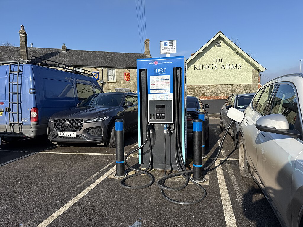 a large blue electric vehicle charging station in a car park with one cable running from it to a silver car. Beyond the car park is a building with “The Kings Arms” painted on it.