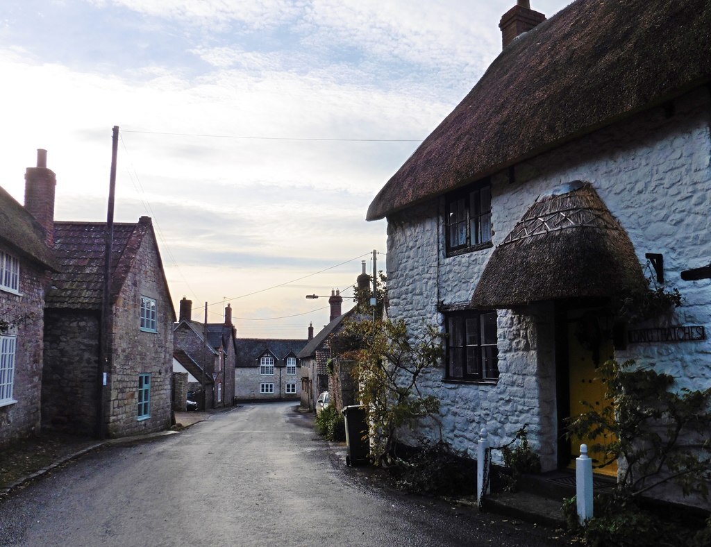 a village road with a white stone cottage with thatched roof and porch on the right and stone buildings along the left. It looks very quaint and traditionally English.