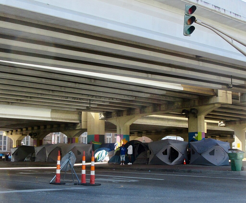 the underpass of a highway with several large tents set up under it. There is an adult and a child standing outside one of the tents.