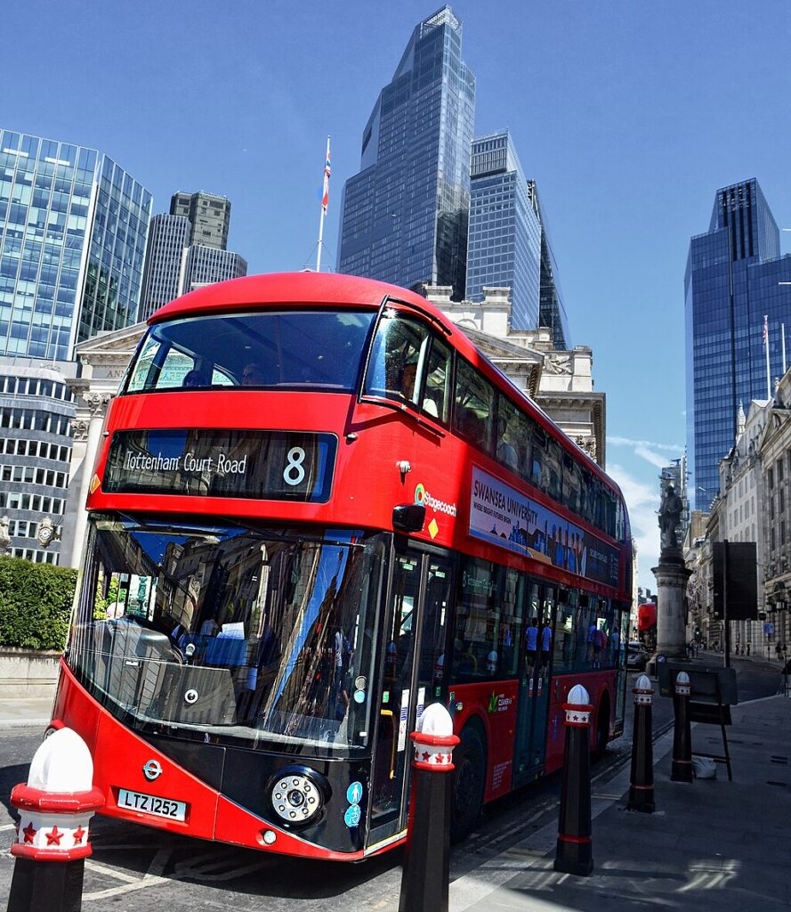 A red number 8 double decker bus driving through London streets. Behind it are skyscrapers reaching up to a clear blue sky.