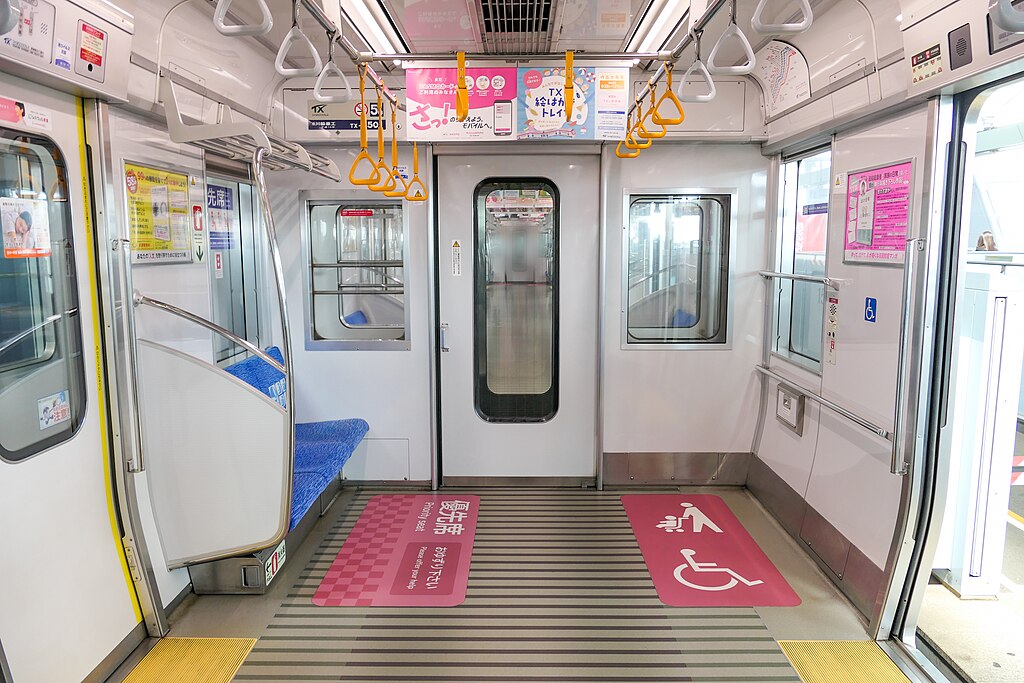 a wheelchair and pushchair space on a train marked with a pink sign on the floor. Above are places for people to grab if standing and across is a seat.