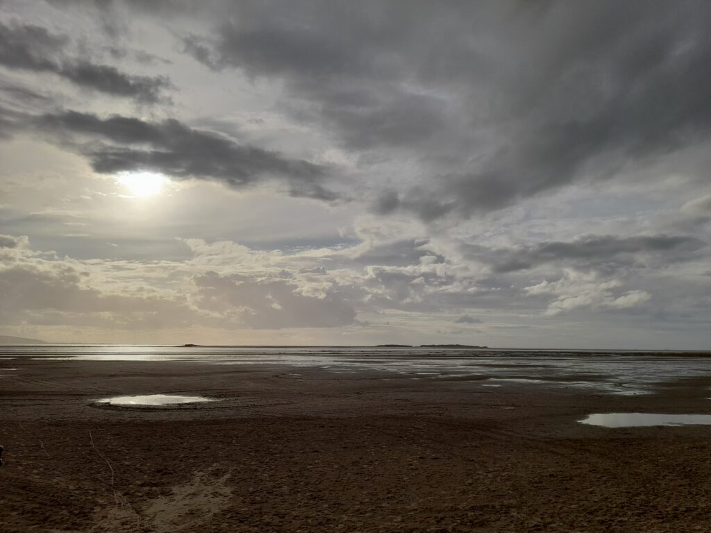 A view of an estuary beach with wide expanse of sky, clouds and sun peeping through
