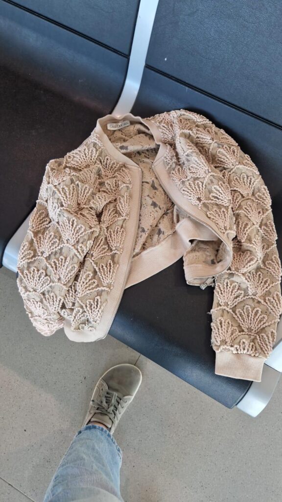 A beige zip cardigan with large crochet flowers in the same colour, on a grey chair.