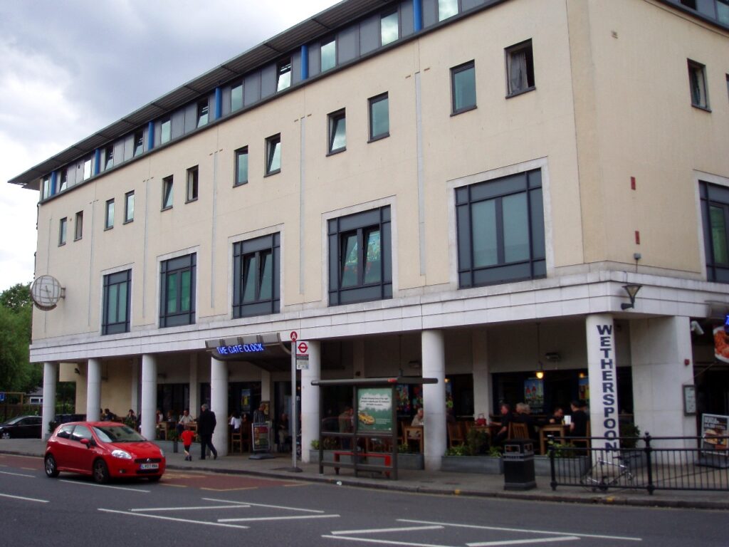 a modern building on a main road with a blue sign for “The Gate Clock” above the entrance and “Wetherspoons” painted on one of the columns holding up the covered area of patio.