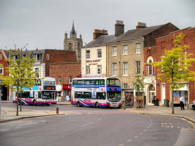 two double decker busses driving through Norwich city centre. There are buildings on one side an what seems to be a pedestrianised area the other side.