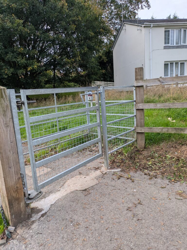 a metal kissing gate between a concrete path and a green grassy area.