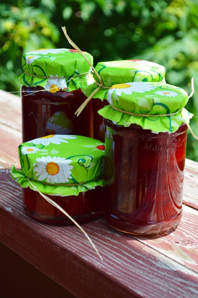 4 different sized pots of red jam with green covers with daisies on them tied on with twine. They rest on a wooden ledge.