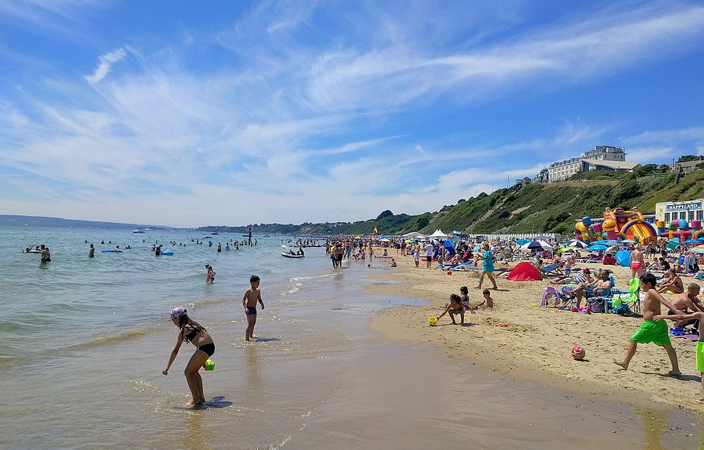Bournemouth beach filled with people playing in the water and along the beach under a bright blue sky with wispy white clouds.