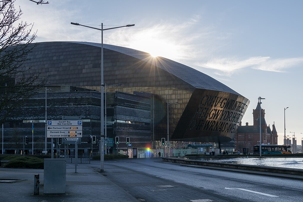The Wales Millennium Centre, a large domed copper coloured building with letters engraved into a section which is leaning over the pathway. The sun is setting behind the building casting large shadows over the road towards it.