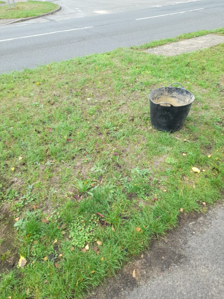a large black bucket with a dirty brown interior sitting on a grassy area beside a road.