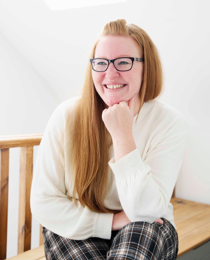 a woman with light skin tone a long straight ginger hair sitting on a wooden bench, one leg crossed over the other, an elbow resting on a leg and her chin resting on a hand. She is smiling.
