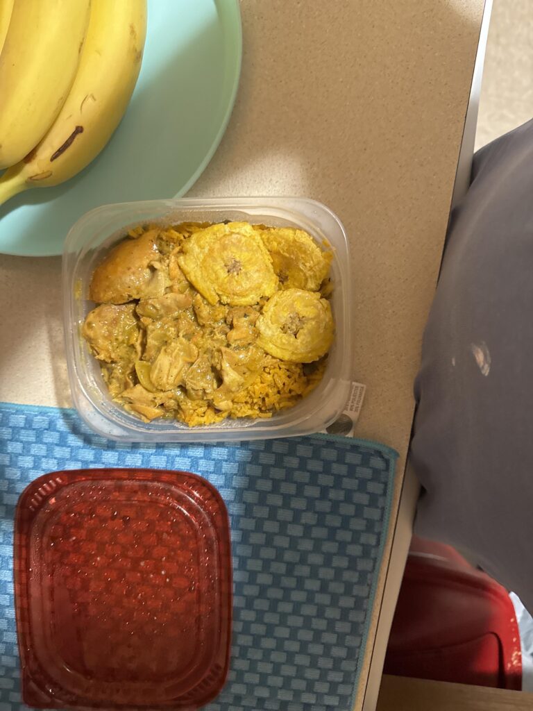 Plastic food container with a meal inside, placed on a counter. The meal includes yellow rice, chicken pieces, and several round fried plantain slices on top, all in a yellow sauce. To the left, there is a light green plate with bananas, and below the container is a woven blue mat with a closed red container on it. Part of someone's clothes and a bit of a red object are visible on the right side of the image. Image description provided with Be My Eyes.