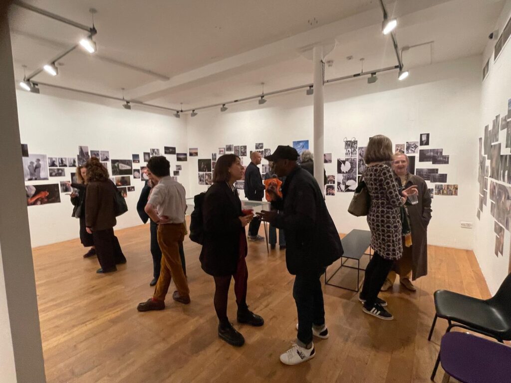 A room with white walls and wooden floor with photos stuck to all the walls. Many people are in the room chatting and laughing and looking at the photography.