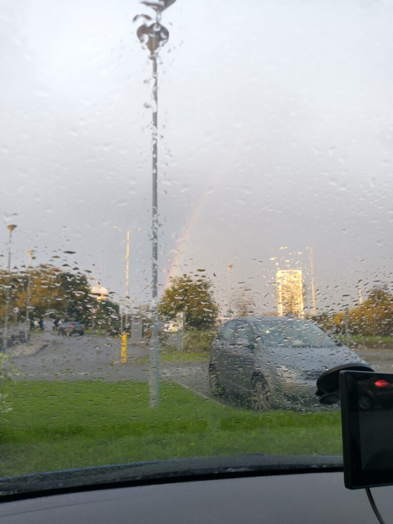 view out of a car windscreen. The windscreen is covered in water, beyond it is a car park with a car in it and a rainbow across the grey sky.