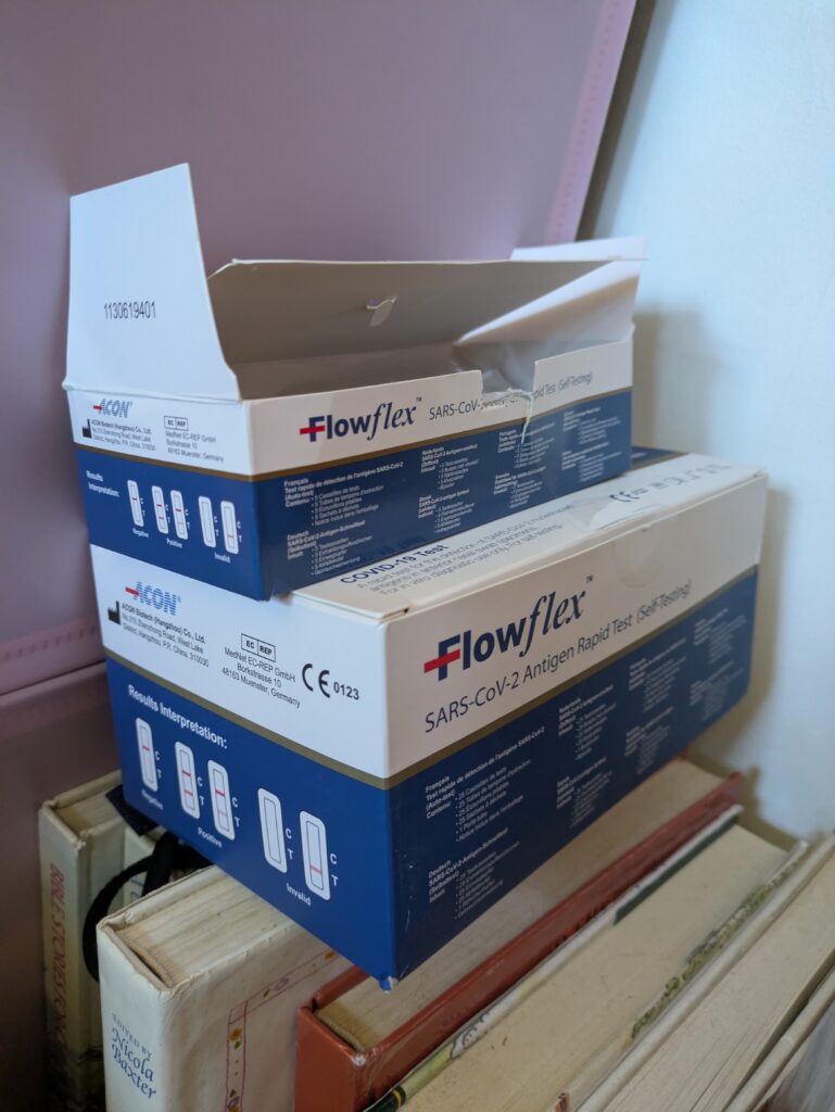 A large box of FlowFlex Sars cov 2 rapid tests and a smaller, open, box of them resting on a stack of books.