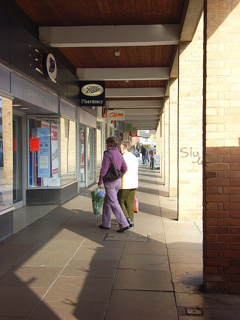 a covered pathway along Woodley shopping precinct with two elderly ladies walking into a Boots, both are carrying bags.