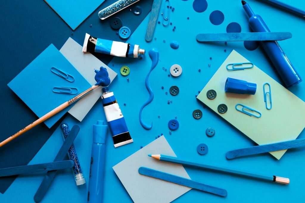 a dark blue table with large blue paper on it covered in blue paint, buttons, pencils, paperclips and other crafting materials.