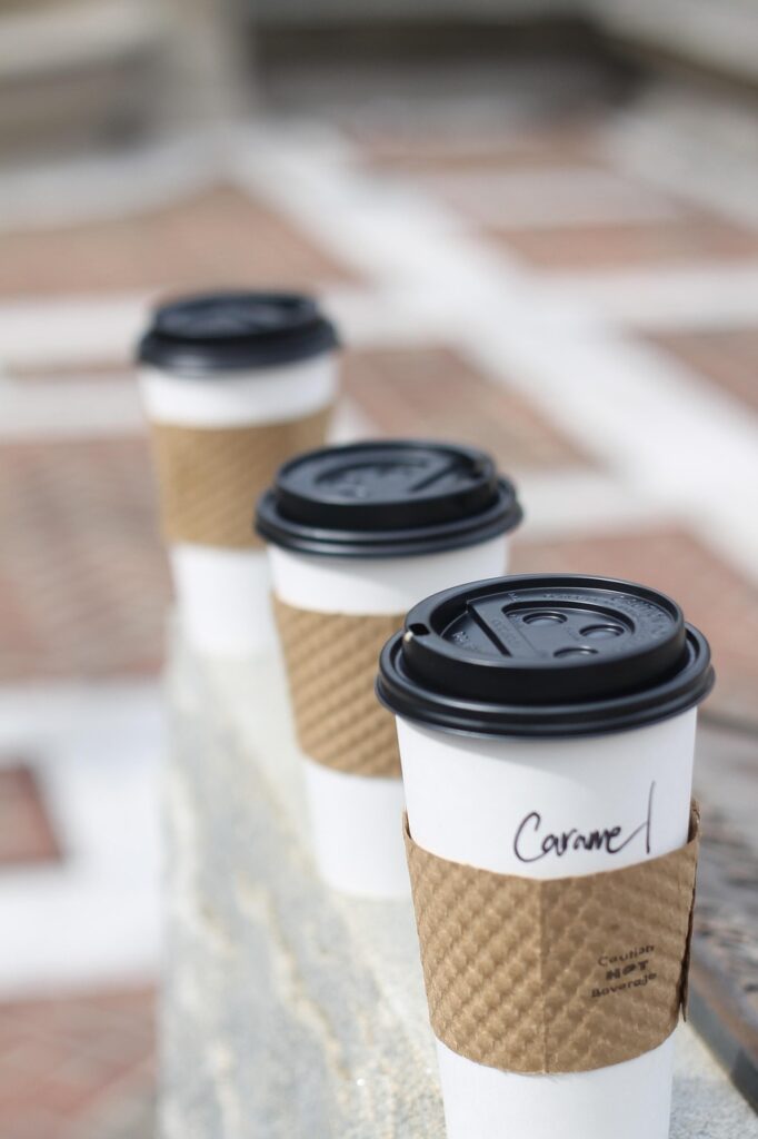 three white takeout coffee cups with brown holder and black lids. The one nearest the camera reads “caramel.”