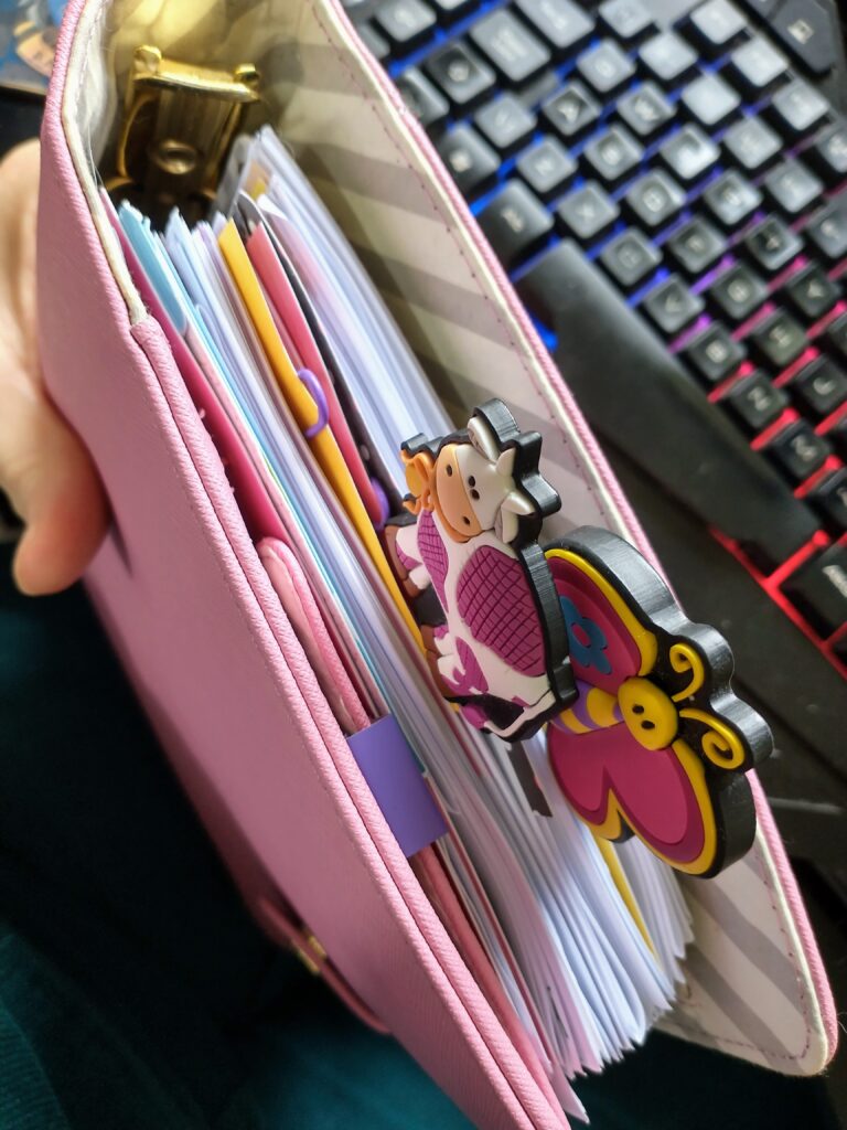 View down into the top of a pink planner filled with paper of different colours. There are two large paper clips one with a silicone cow and one a butterfly on them sticking out. A hand is holding the planner.