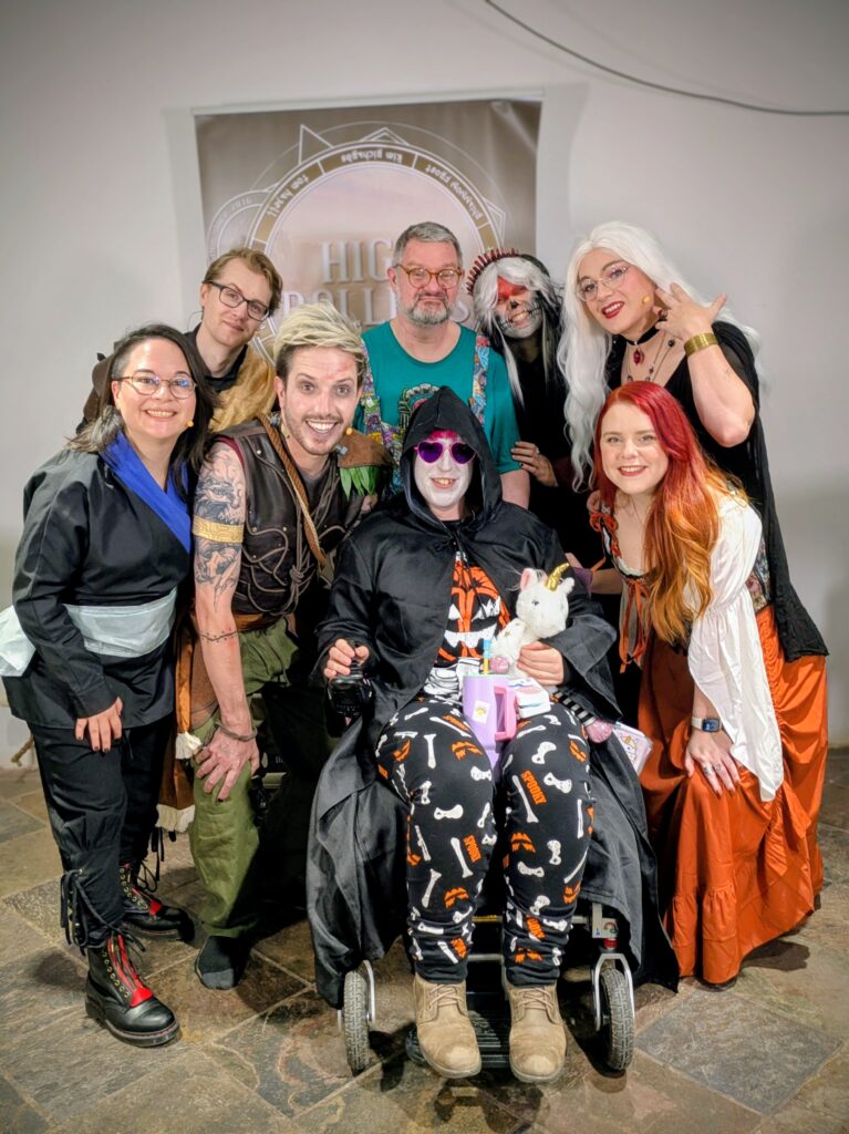 A group of people dressed up in fantasy and halloween outfits. A woman in the front is a wheelchair wearing leggings with bones on and a top with a skeleton and pumpkin on it, a black cape with hood and her face painted white wearing heart shaped glasses with purple lenses and pink frames. A man is stood behind her in a green t-shirt and dungarees. Around them are arranged the High Rollers cast dressed as their characters and Mara in a fancy black dress looking very glamorous.
