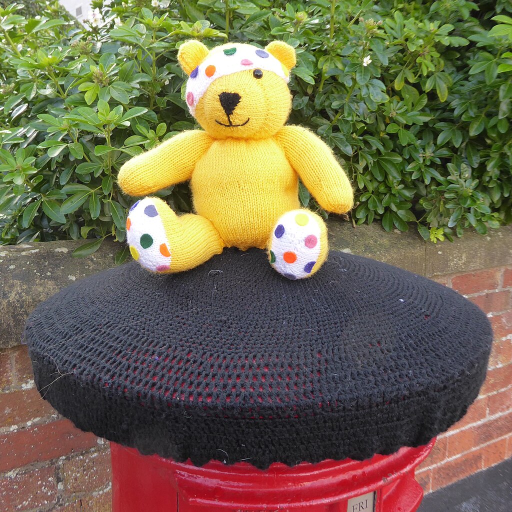a knitted Pudsey bear, mascot for Children In Need, wearing the iconic spotted eye patch, sitting on a black knitted top which covers a red post box.