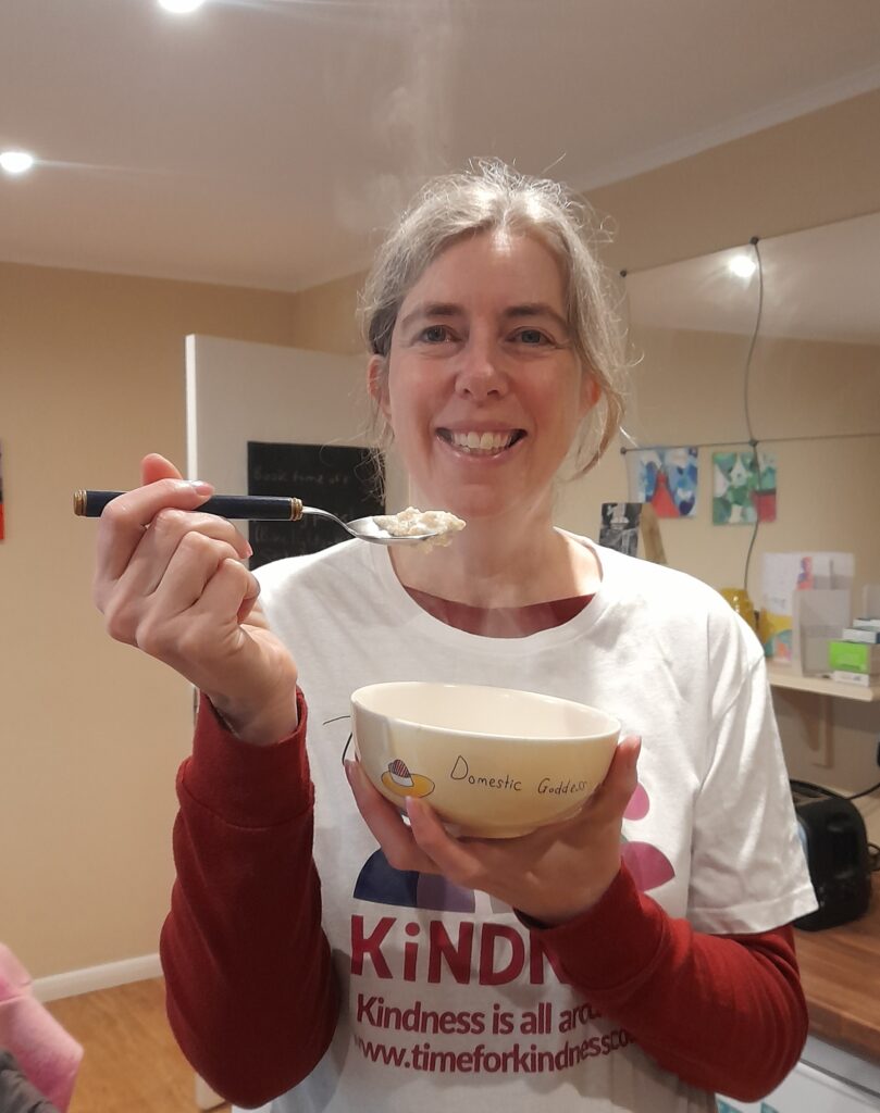 Sarah, a woman with light skin tone and long grey hair tied back, wearing a white t-shirt with the Time for Kindness logo on it over a long sleeved red top. She is holding a bowl of steaming hot porridge and a spoon with some on it above and smiling.