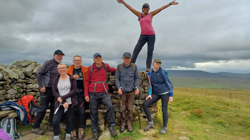 A group of 7 people dressed in walking gear stand outdoors in front of a beautiful view in the Yorkshire Dales - 1 of them is standing on top of a stone wall with her arms thrown joyfully into the air