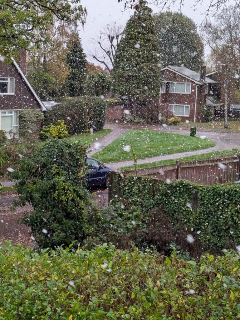 view through a window onto a housing estate as a car drives past and snow falls from the grey sky.