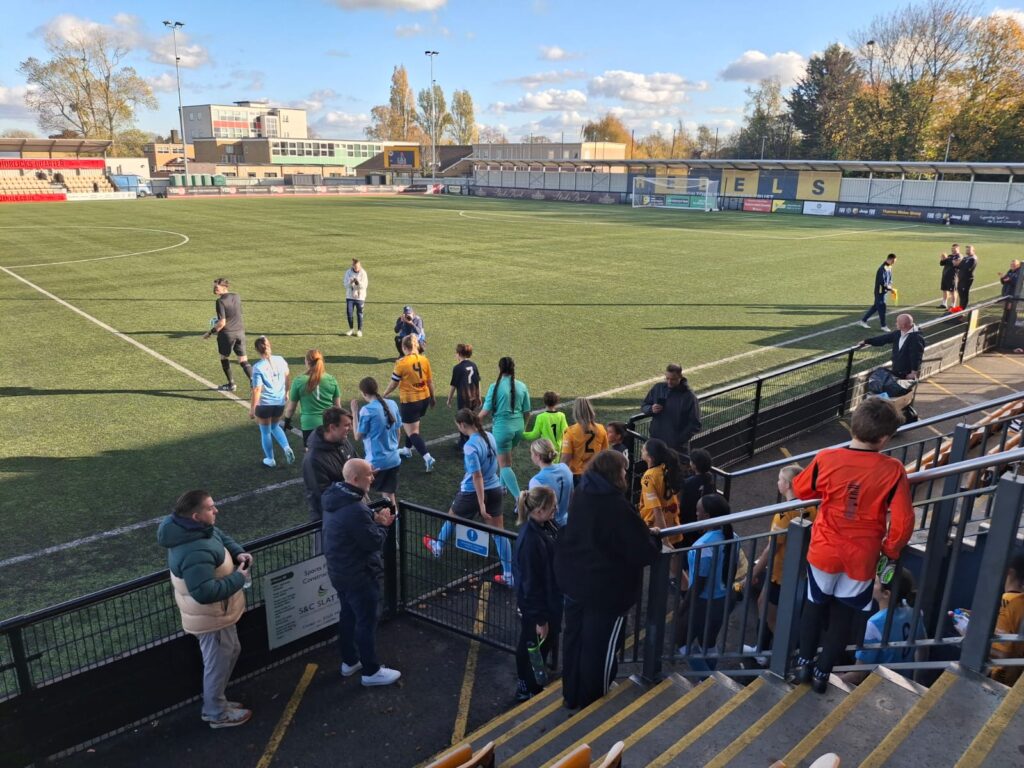 a women’s football club walking out onto the pitch on a sunny day. The team are wearing a light blue kit.