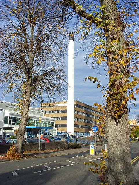 In the foreground is a tree beside a road. Beyond which is a medium sized white building with a lot of cars parked outside and next to it a 4 story squarish building with a large white chimney on the corner of it more than twice as tall as the building.